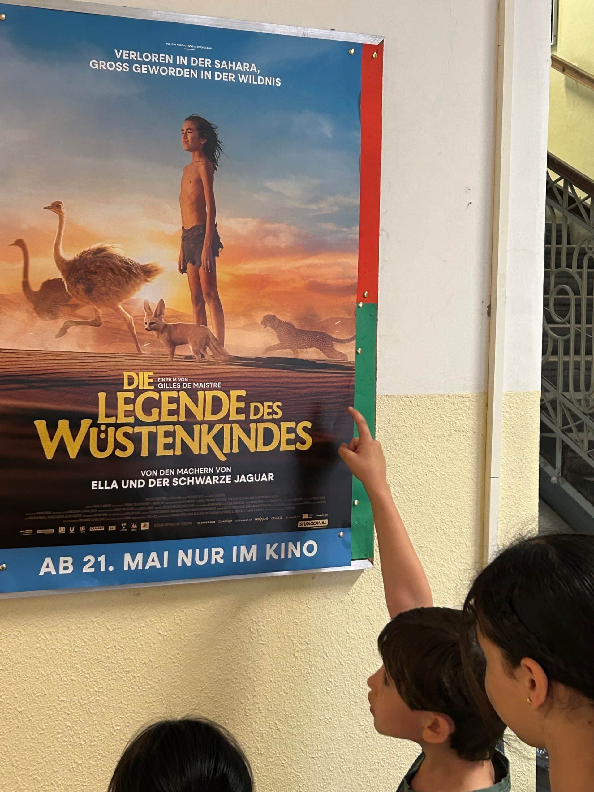 Drei Kinder betrachten ein Filmplakat für „Die Legende des Wüstenkindes“, auf dem ein Junge, Strauße und eine Landschaft zu sehen sind, und zeigen darauf; das Plakat hängt an einer Wand.
