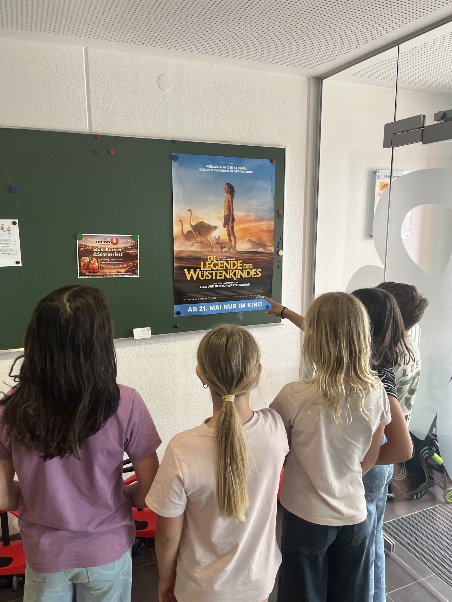 Vier Kinder stehen vor einer Pinnwand und betrachten ein Plakat zum Film „Die Legende des Wüstenkindes“; eines der Kinder zeigt auf das Plakat.