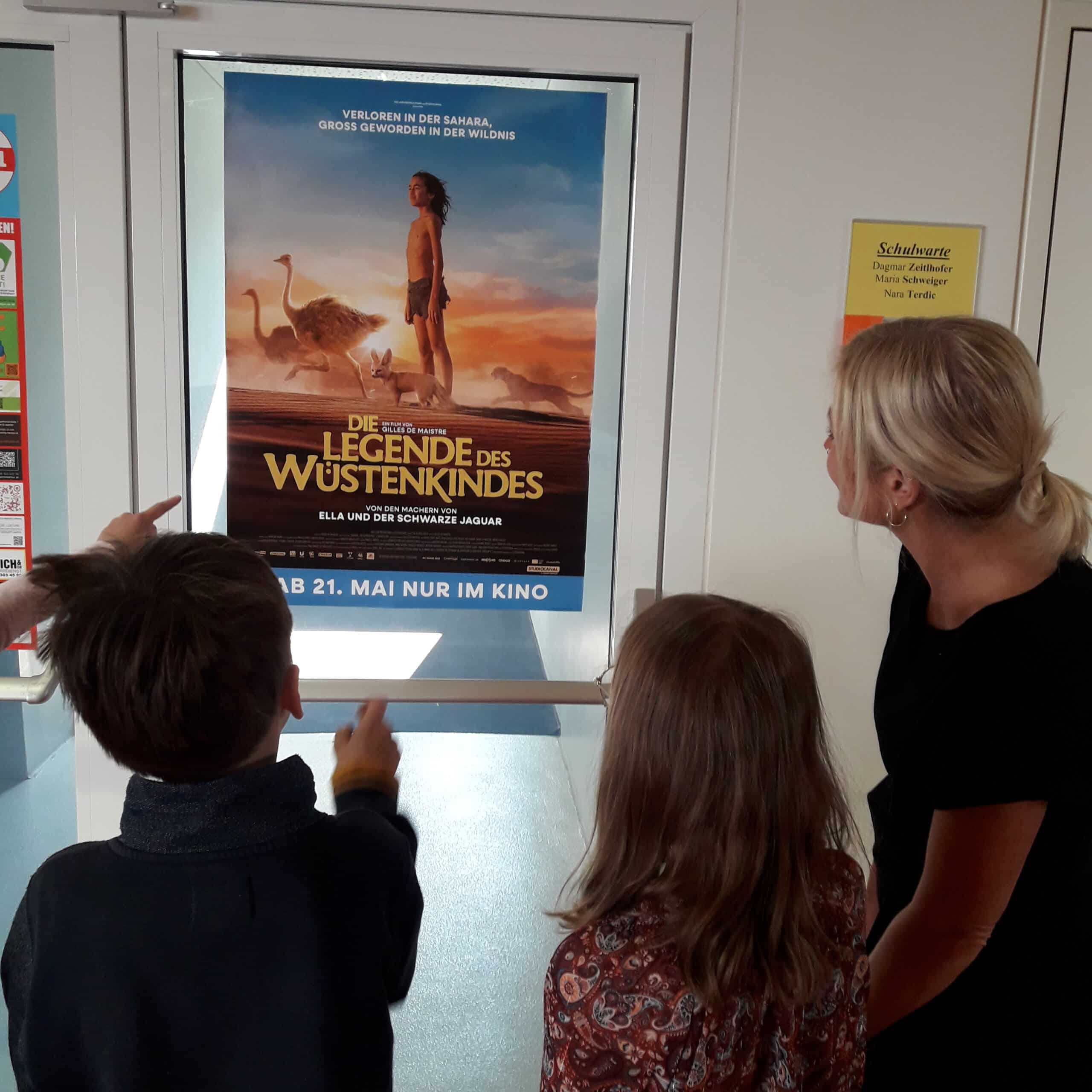 Zwei Kinder und eine Frau betrachten ein Filmplakat zu „Die Legende des Wüstenkindes“, auf dem eine Person mit Straußen in der Wüste zu sehen ist.