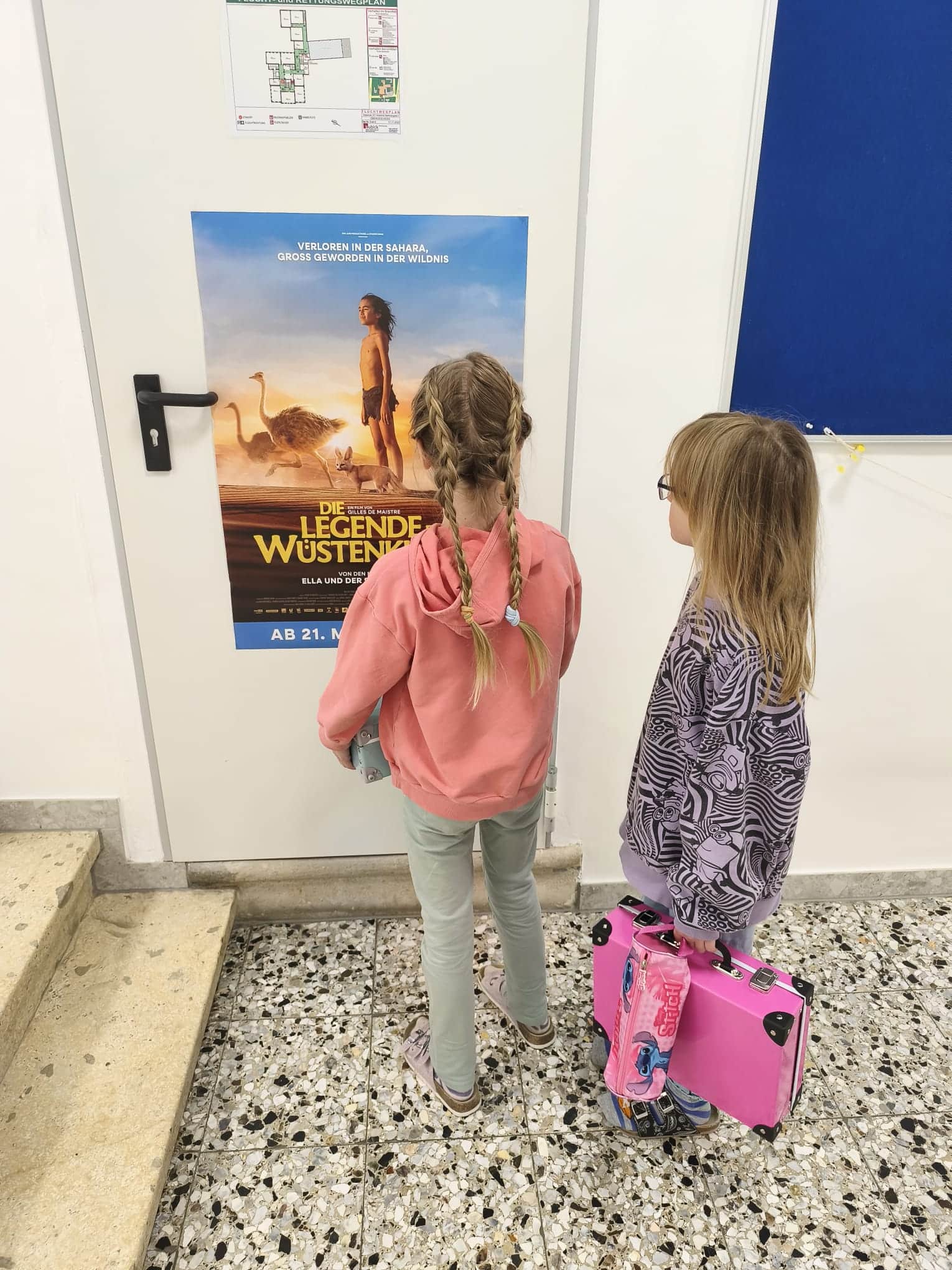 Zwei junge Mädchen stehen vor einer Wand und betrachten ein Filmplakat für „Die Legende der Wüstenkönige“. Eines der Mädchen hält einen rosa Koffer in der Hand.