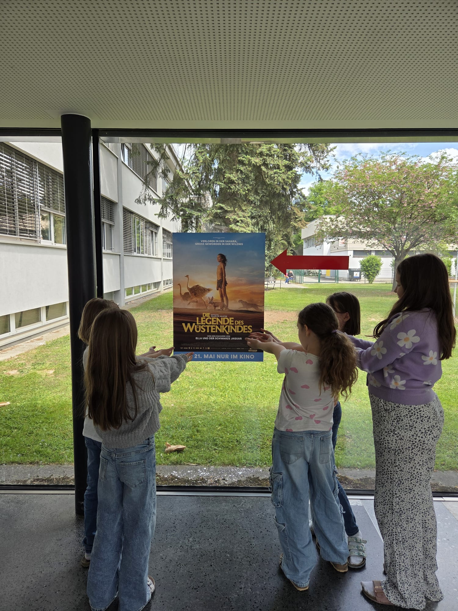 Vier Kinder stehen im Innenraum und zeigen jeweils auf ein Filmplakat für „Legende der Wüstenkinder“, das an einer Glastür angebracht ist, die auf einen grasbewachsenen Außenbereich hinausgeht.