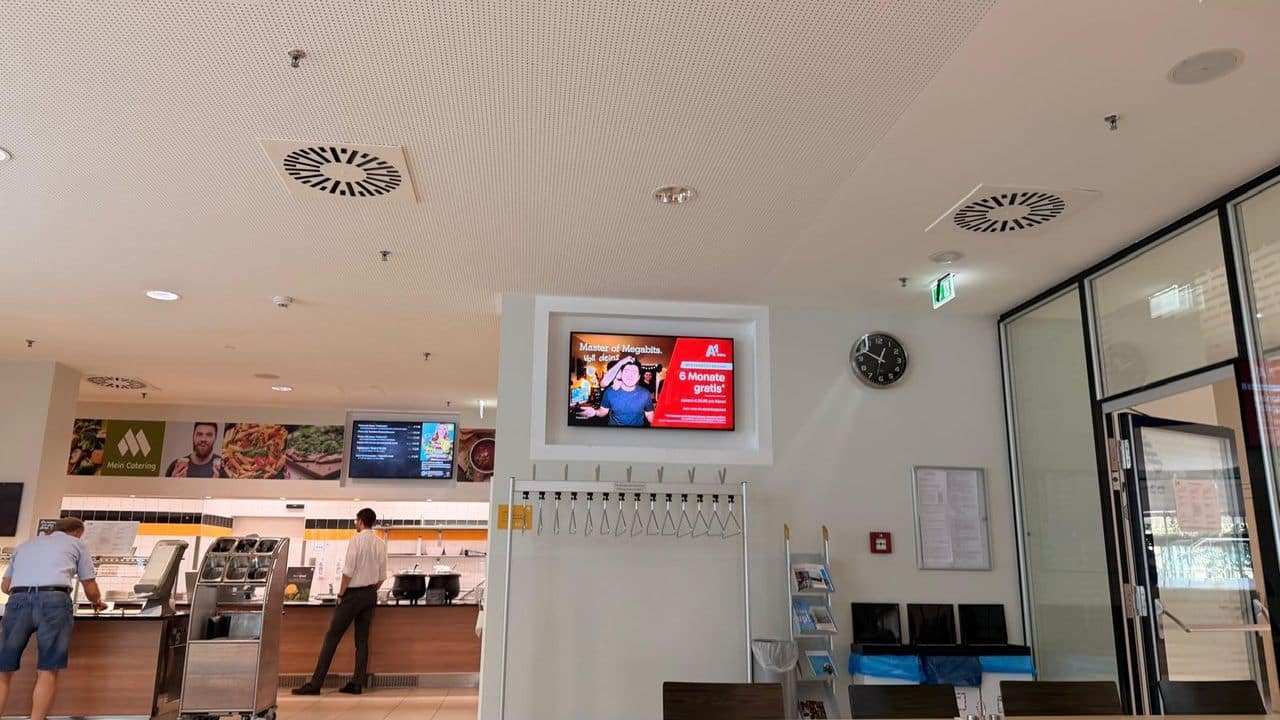 Eine Cafeteria mit einem digitalen Bildschirm, auf dem eine Werbung läuft, einer Uhr an der Wand, Kleiderhaken und zwei Personen in der Nähe der Essensausgabe.