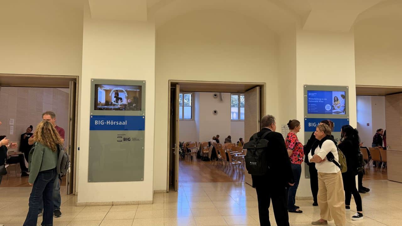 Vor zwei großen Hörsälen, in denen andere sitzen, versammeln sich Menschen im Flur und unterhalten sich. Auf Schildern neben den Türen steht „BIG-Hörsaal“.