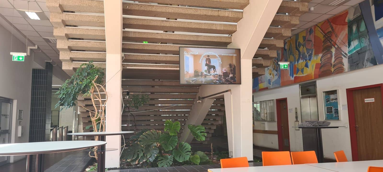 Ein Innenraum mit einer Treppe, einem Fernseher, auf dem eine Präsentation läuft, mehreren Pflanzen, bunten Wandmalereien und orangefarbenen Stühlen, die um weiße Tische herum stehen.