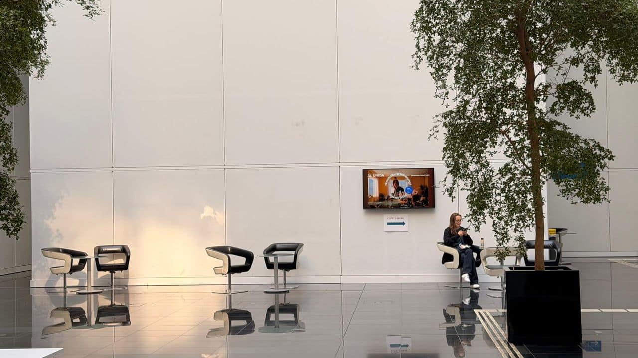 Eine Person sitzt allein auf einem schwarzen Stuhl in einer modernen, geräumigen Lobby mit großen Topfbäumen, einem Fernseher an der Wand und mehreren leeren Stühlen.