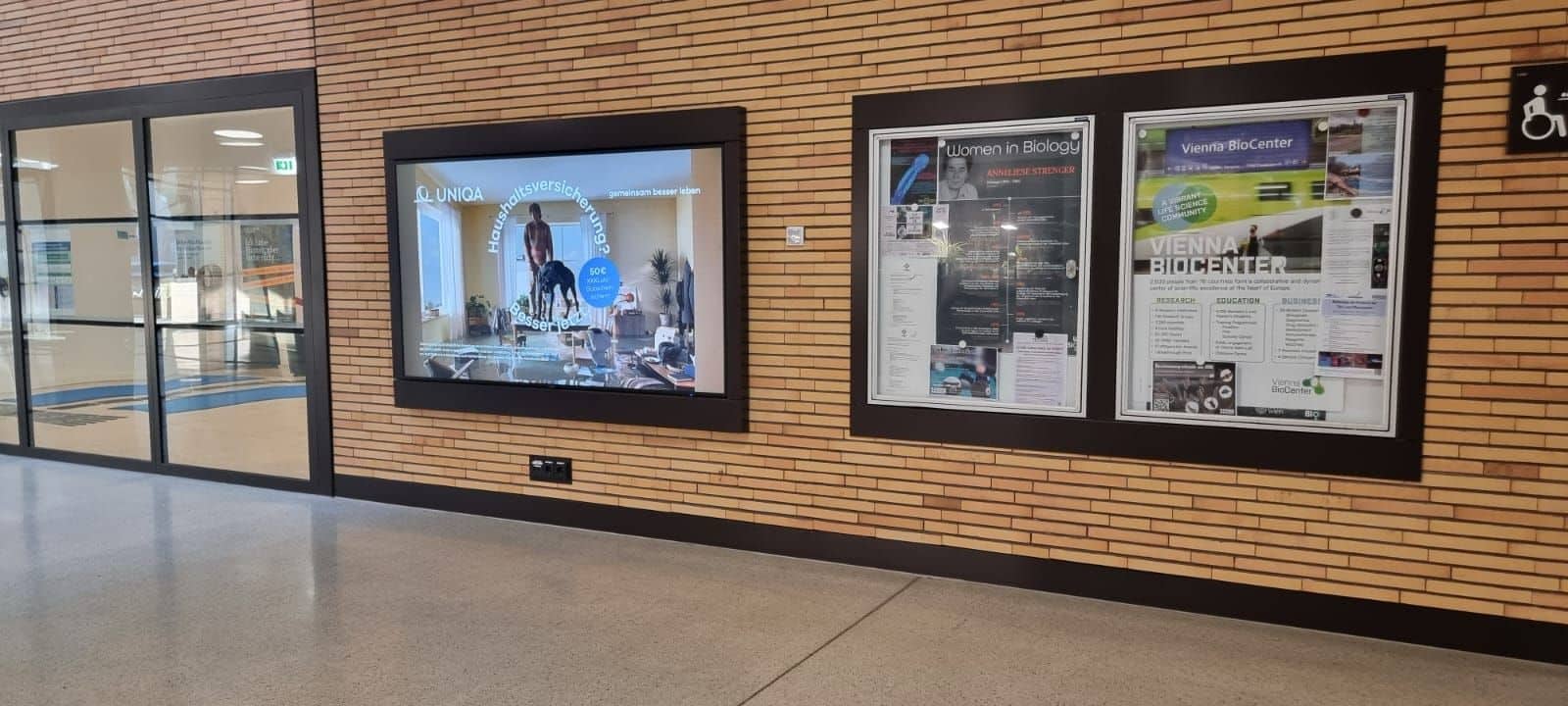 Ein Flur mit einer digitalen Werbeanzeige an der linken Wand und einer Pinnwand mit Plakaten und Flyern auf der rechten Seite. Der Boden ist poliert und die Wand besteht aus Ziegelsteinen.