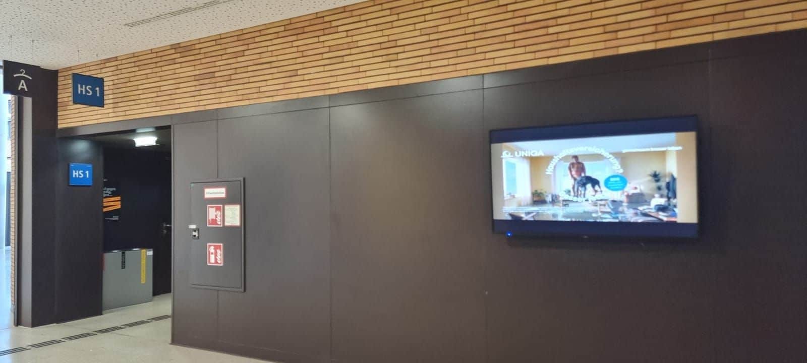 Ein Flur mit einem blauen Schild mit der Aufschrift „HS1“, einer Brandschutztafel, einem Aufzug und einem an der Wand montierten digitalen Bildschirm, auf dem eine Werbung angezeigt wird.