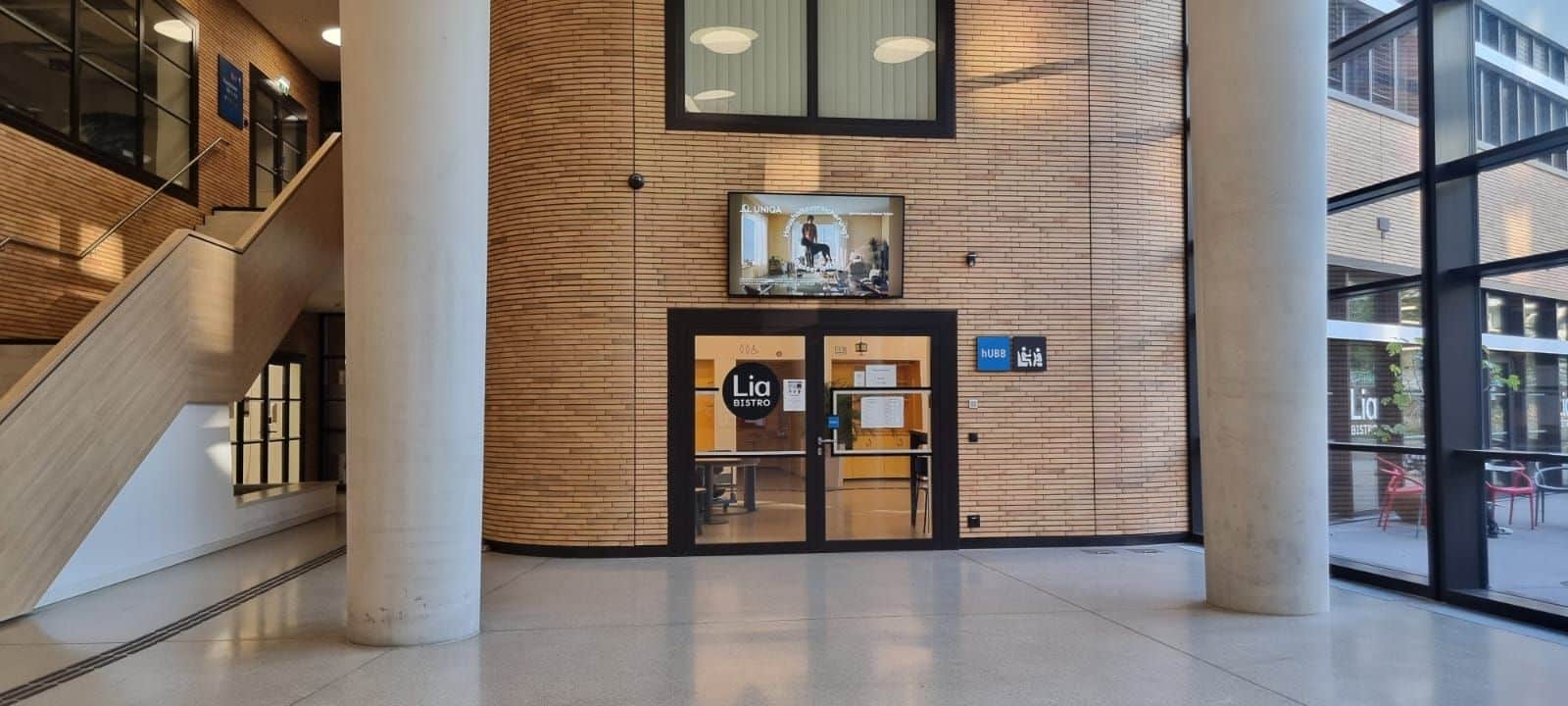 Eingang zu einem Raum mit der Aufschrift „Lia Retro“, mit Glastüren, einem Schild, einem darüber hängenden Fernsehbildschirm und einer Treppe auf der linken Seite in einem modernen Gebäude mit Backsteinwänden und großen Fenstern.