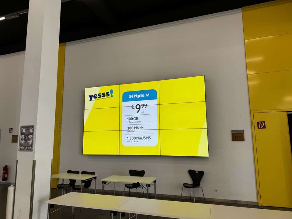 Auf einem großen digitalen Bildschirm wird ein „yesss!“-Mobilfunktarif angezeigt: 9,99 €, 100 GB Datenvolumen, 200 Mbit/s, 1500 Min./SMS. Darunter stehen in einem weiß-gelben Raum Tische und Stühle.
