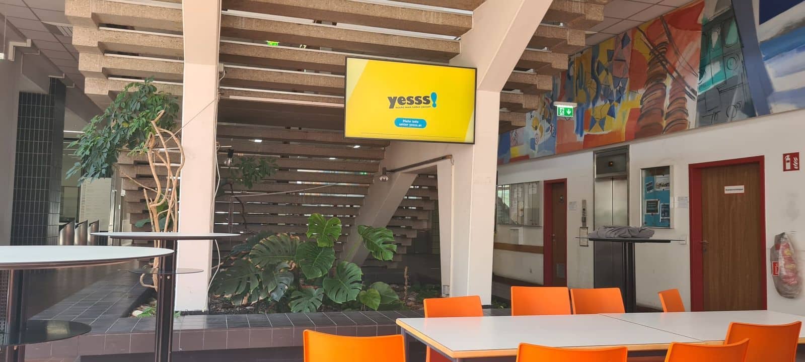 Ein Cafeteria-Bereich mit orangefarbenen Stühlen, weißen Tischen, einer Treppe, Pflanzen, einem Wandbild und einem Bildschirm, auf dem „yesss!“ und „Wir sind im WLAN“ zu lesen ist.