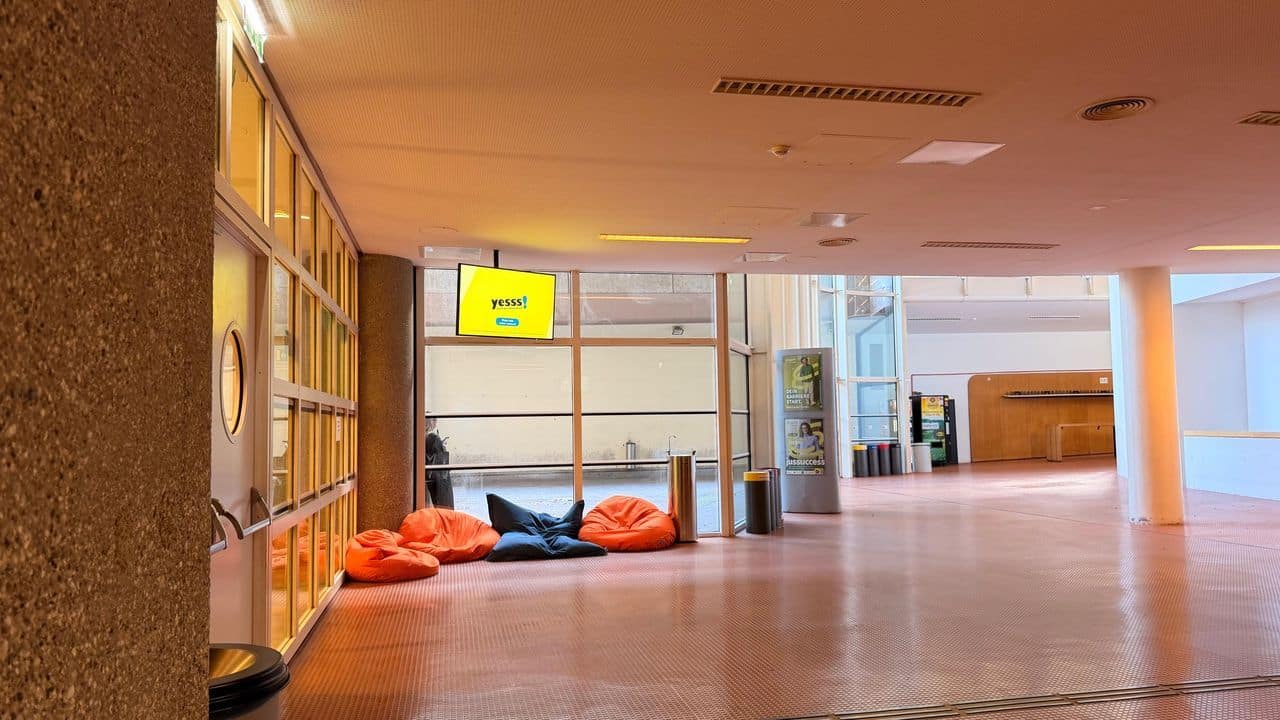 Ein Flur mit orangefarbenen und blauen Sitzsäcken in der Nähe von Glastüren, einer gelben Digitalanzeige über den Köpfen und einem Automaten im Hintergrund.