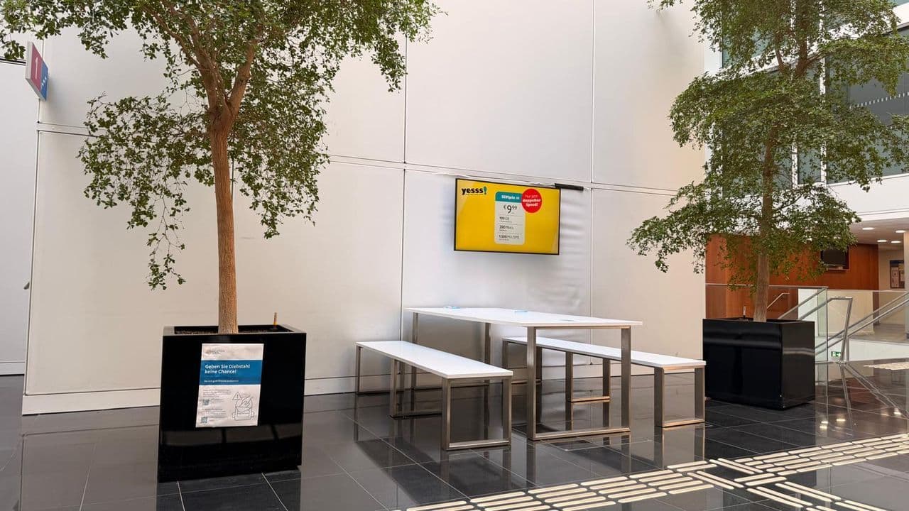 In einem modernen Innenraum stehen weiße Sitzbänke und Tische zwischen zwei großen Topfbäumen, während an der weißen Wand dahinter ein digitaler Bildschirm Informationen anzeigt.