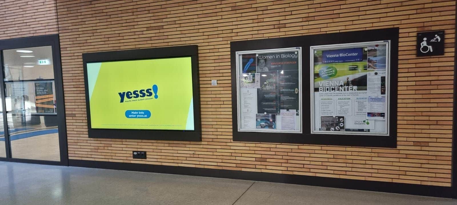 An einer Backsteinwand in einem Flur ist neben zwei Anschlagtafeln mit Plakaten und Aushängen ein digitaler Bildschirm zu sehen, auf dem eine gelbe „yesss!“-Werbung angezeigt wird.