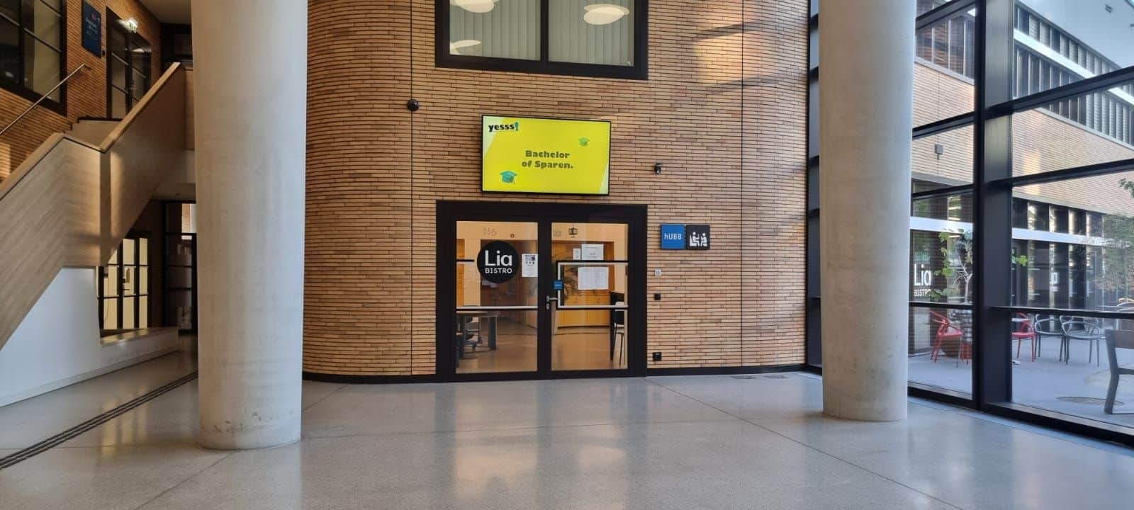 Ein Flur mit einem gläsernen Eingang zu einem Raum mit der Aufschrift „Lia Bistro“. Über der Tür befindet sich ein gelbes Schild mit der Aufschrift „Hostelería y Oportun“. Die Toiletten sind durch Schilder auf der rechten Seite gekennzeichnet.