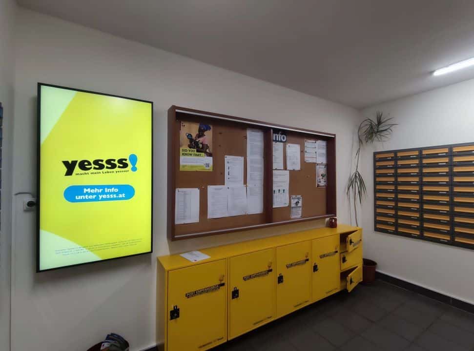 Ein Flur mit gelben Spinden, einer mit Zetteln übersäten Pinnwand, einer Reihe von Briefkästen und einer leuchtend gelben „yesss!“-Werbung an der Wand.