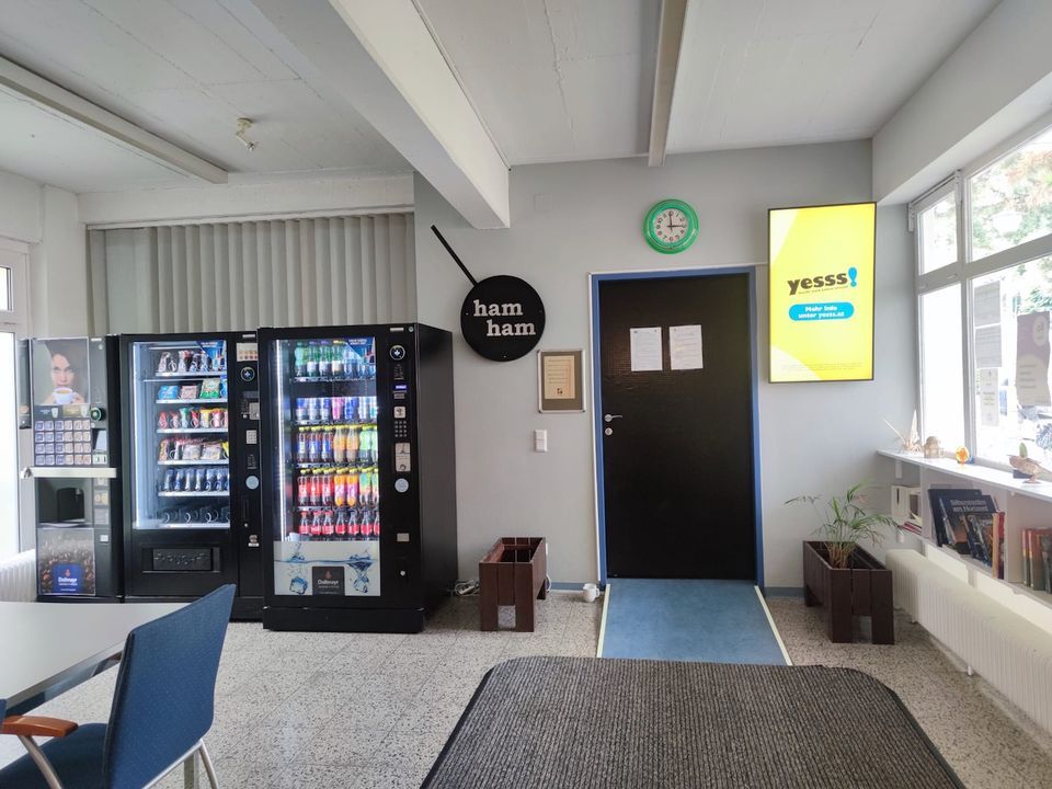 Zwei Verkaufsautomaten und eine Recyclingstation stehen an einer Wand in einem hell beleuchteten Raum, in dem eine Uhr, ein Digitalbildschirm und ein Schild mit der Aufschrift „Ham Ham“ zu sehen sind.