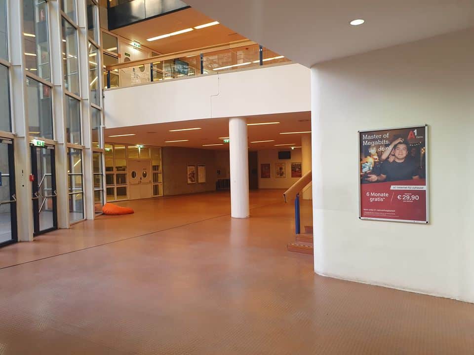 Ein geräumiger Flur mit großen Fenstern, einigen Postern an den Wänden, einem orangefarbenen Sitzsack, einer Treppe und einer Anzeige für eine Zeitschrift, die auf einer weißen Säule zu sehen ist.
