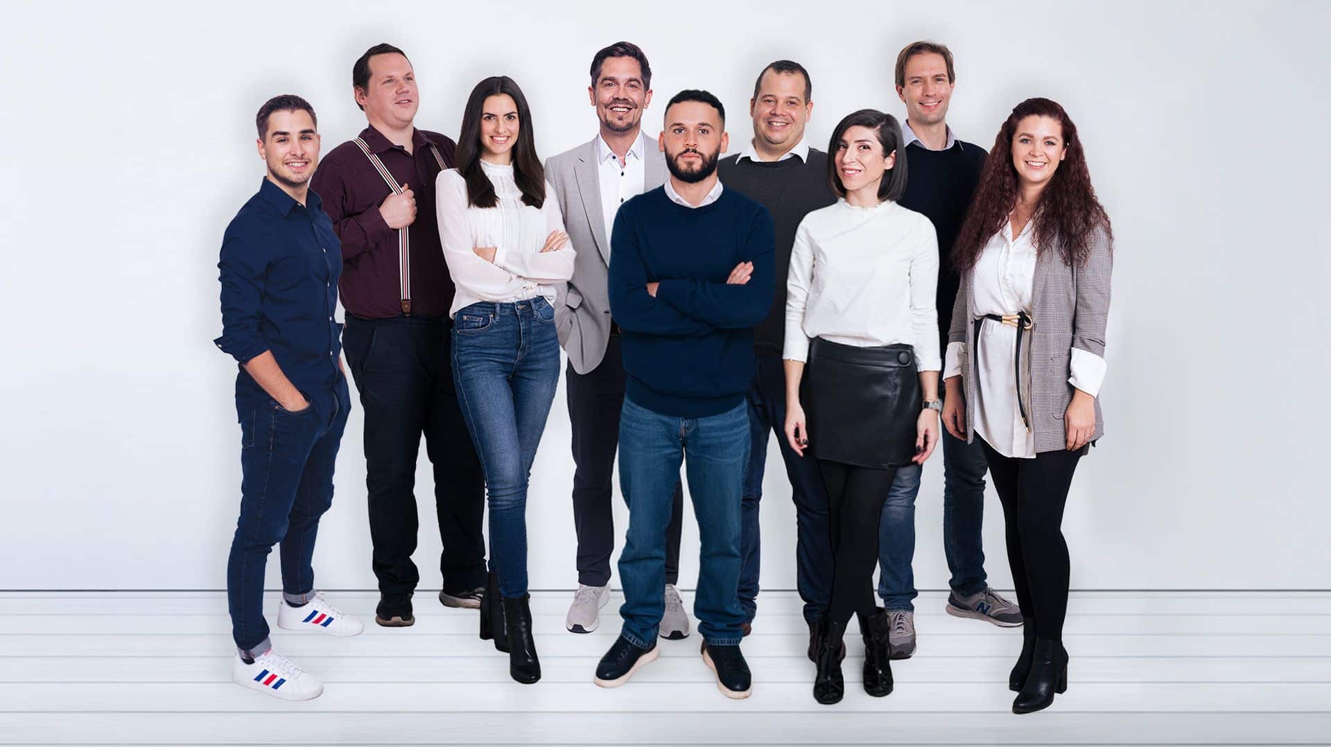 Neun Erwachsene stehen gemeinsam vor einem weißen Hintergrund, gekleidet in Freizeit- und Business-Casual-Kleidung, und posieren lächelnd für ein Teamfoto der Young Enterprises Media GmbH